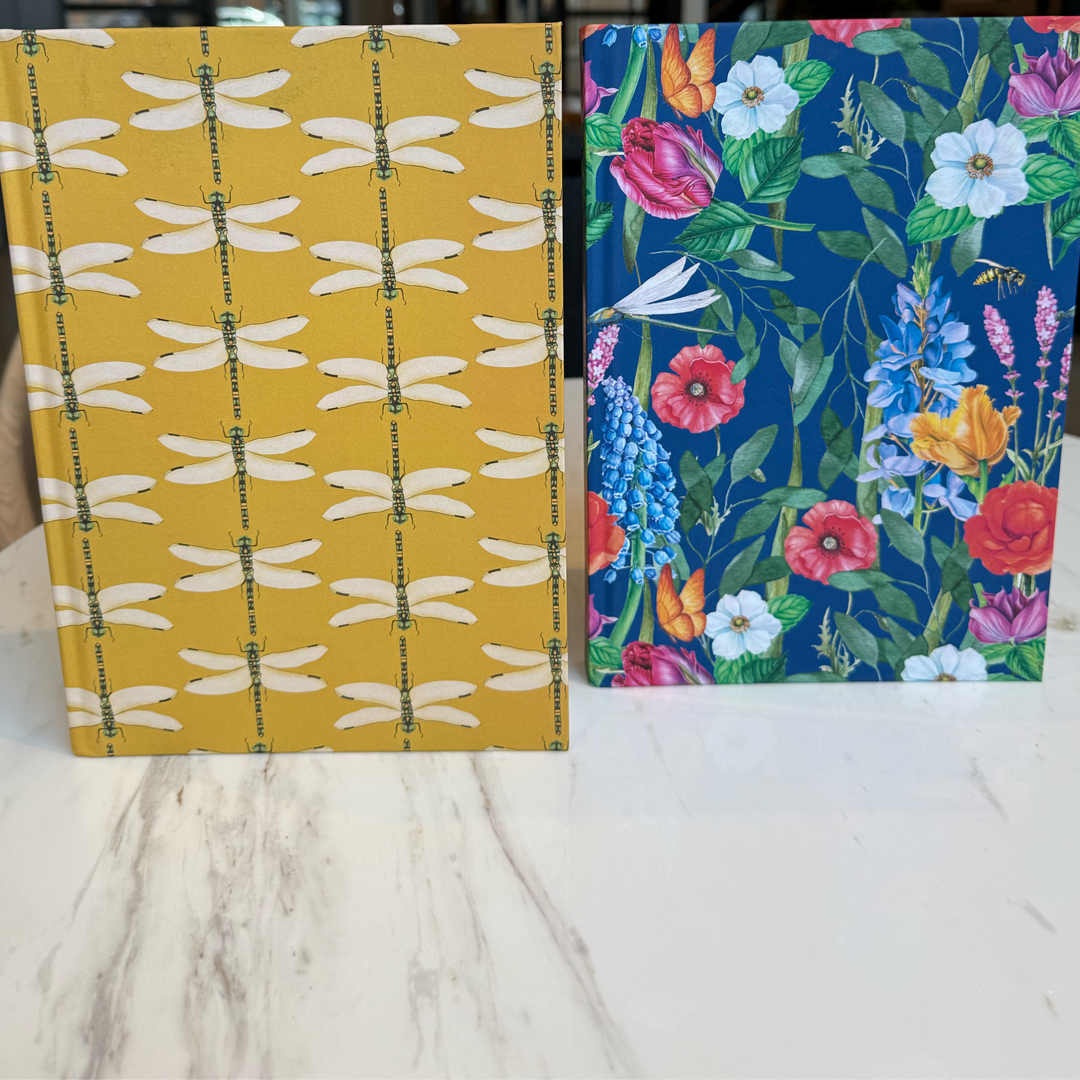 Two journals and planners with different covers, one chartreuse with dragonfly pattern and the other blue with floral pattern, on a marble desk.