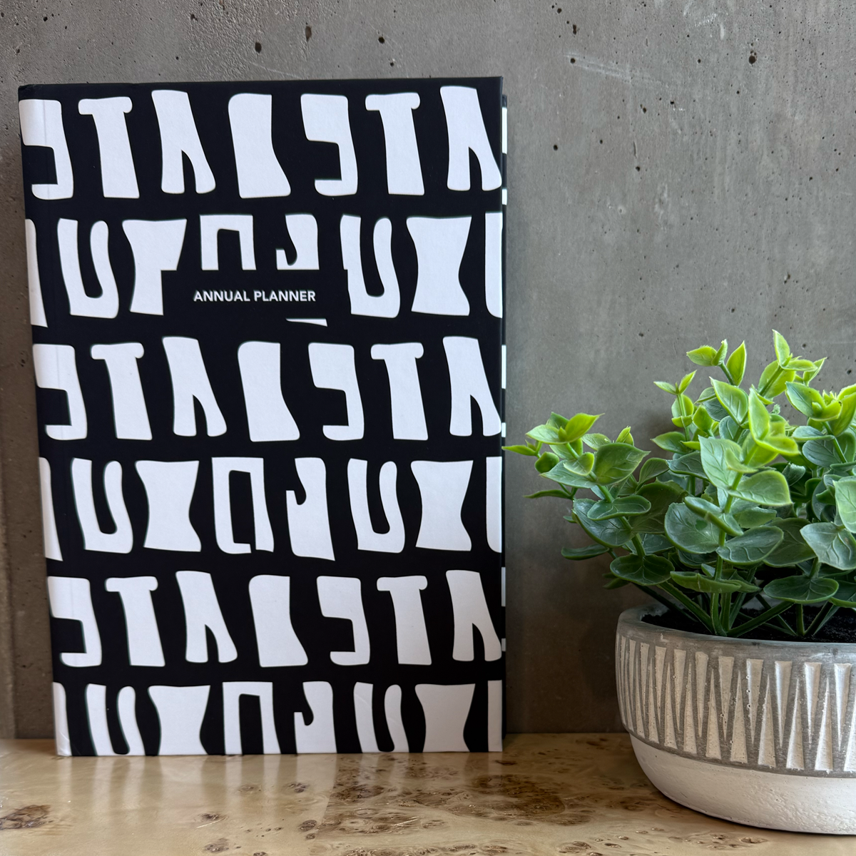 Black and white planner with batik pattern next to a potted plant on a wooden desk.