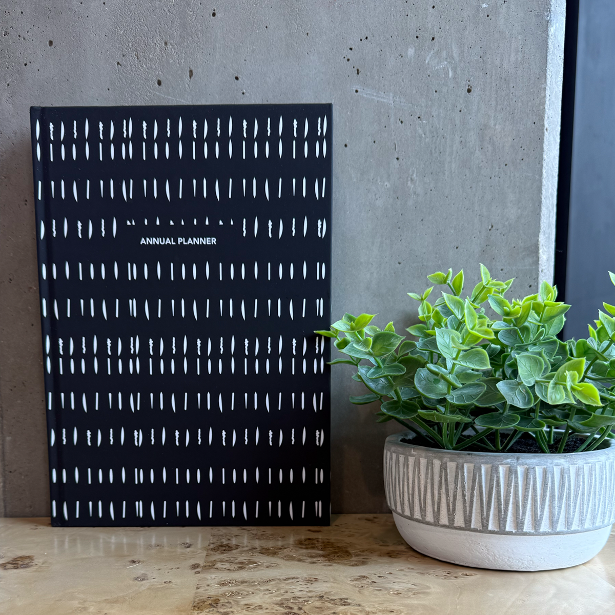 Black planner with white patterns and a potted plant on a wooden desk.